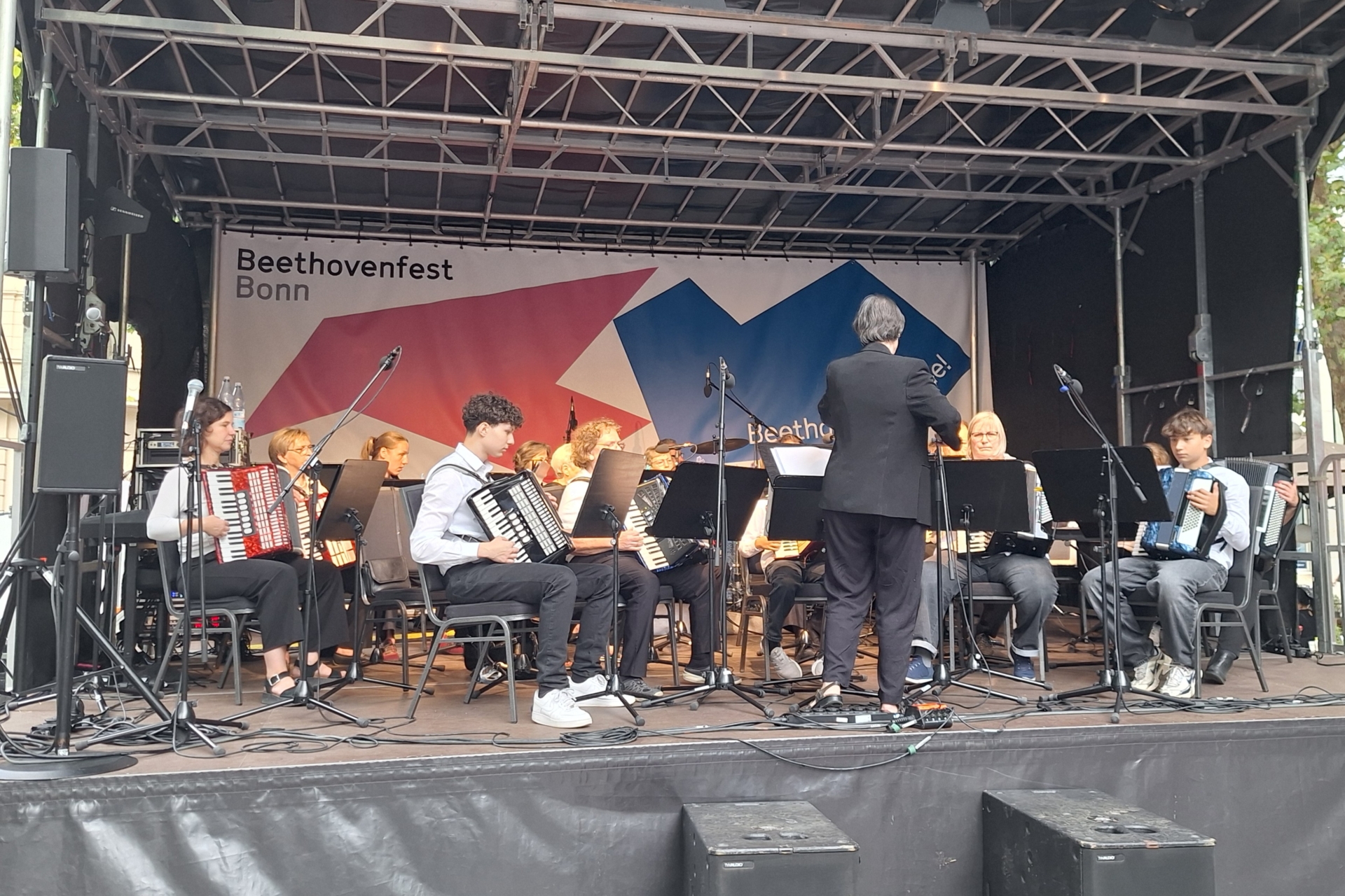 Auf einer tiefen Bühne sitzen die 18 Akkordeonistinnen und Akkordeonisten inklusive Schlagzeug, Bass und Keyboard beim Konzert.