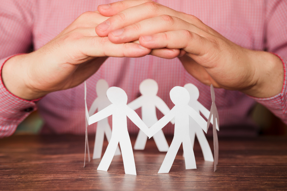 Schützende Hände über einem Kreis aus Papier-Figuren die Menschen darstellen. Sinnbildlich für Versicherungsschutz.