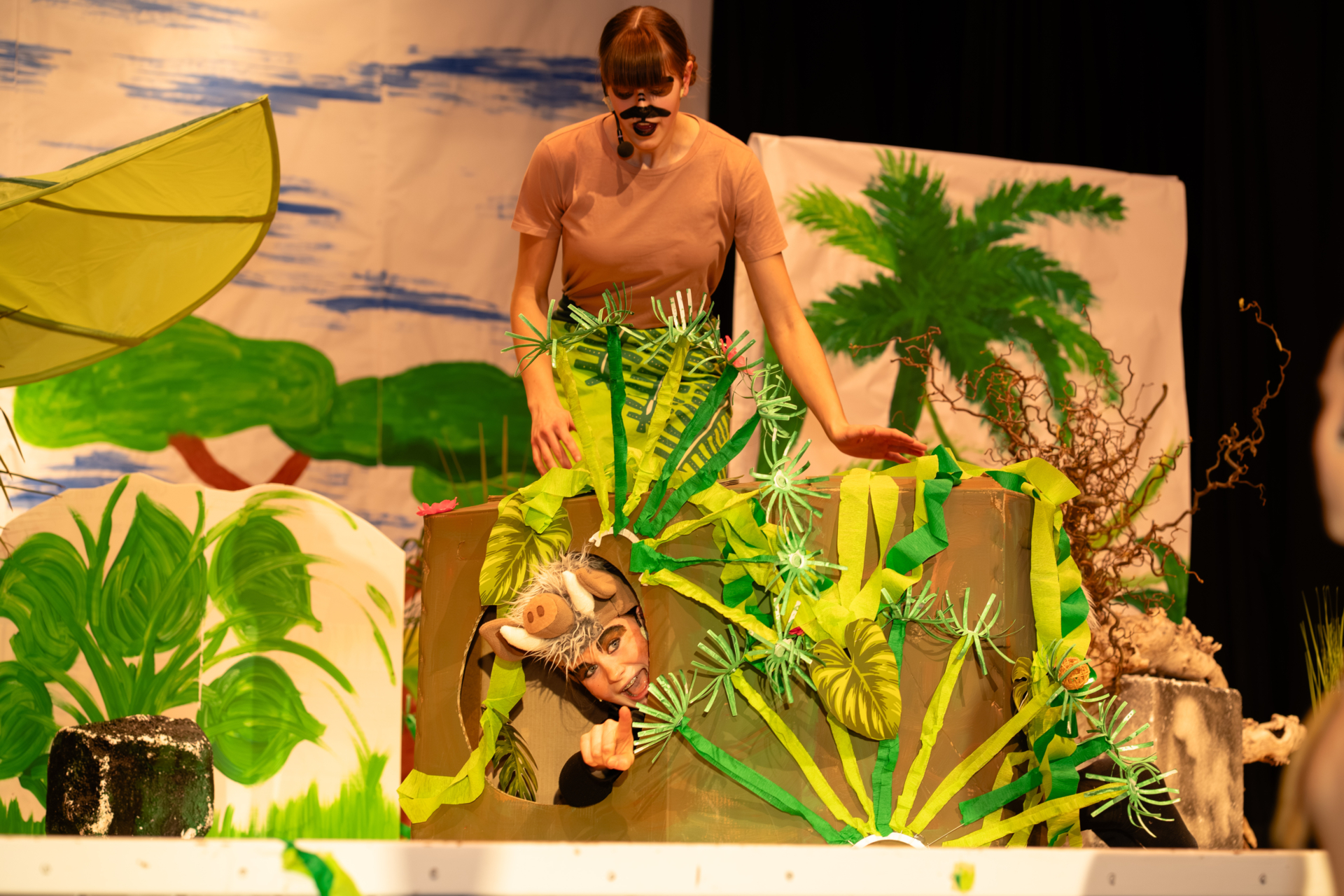 Timon und Pumbaa während einer Szene: Zu sehen sind die beiden Schauspieler auf der Bühne mit sehr viel hauptsächlich in grün gehaltener Kulisse. Pumba versteckt sich gerade in einem Karton, der eine Erdhöhle darstellen soll und schaut dort heraus. Er zeigt mit dem Finger in Richtung der drohenden Gefahr.