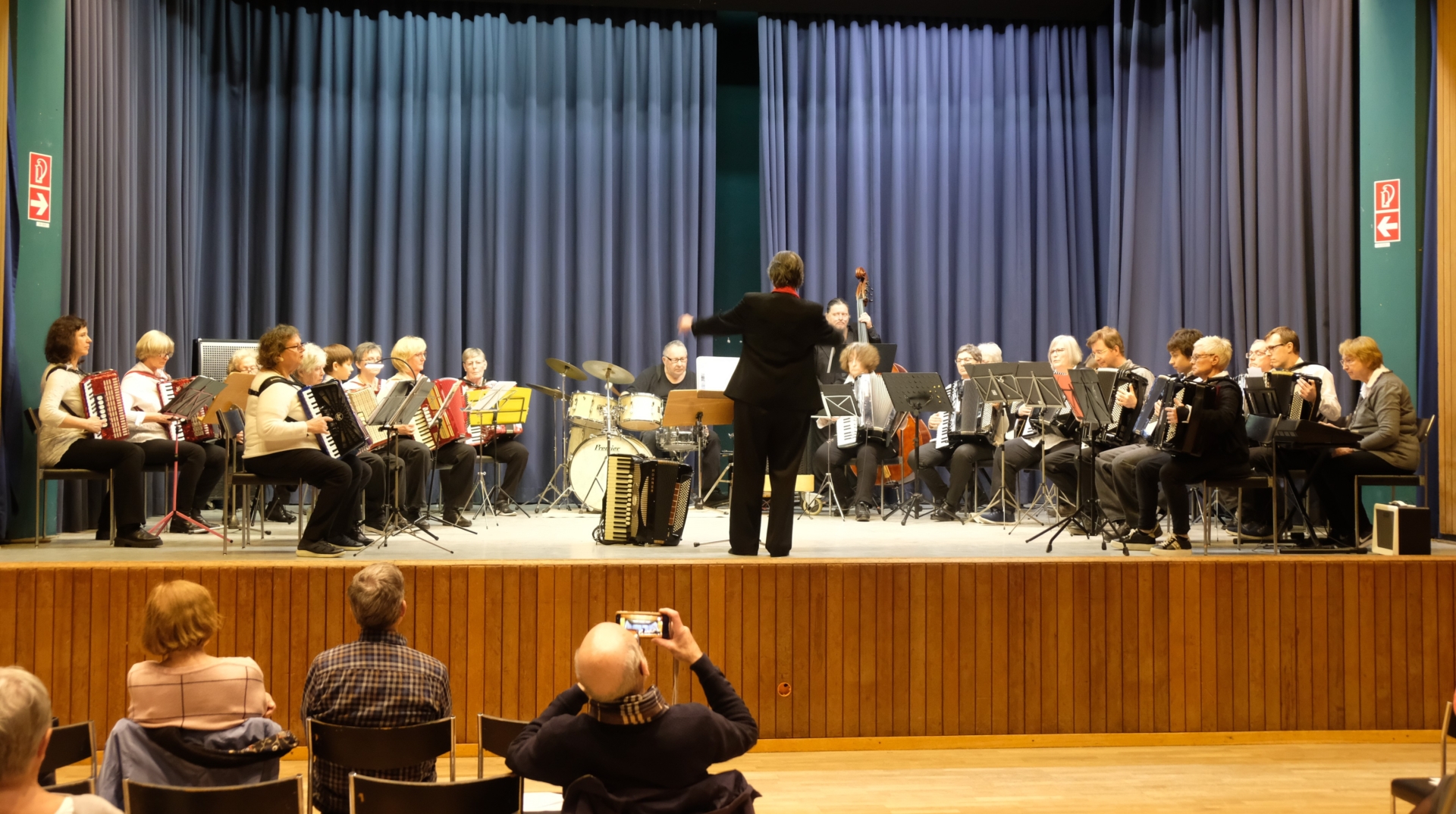 Blick auf die Bühne mit Dirigentin und dahinter den 23 Aktiven des Orchesters verschiedenen Alters.