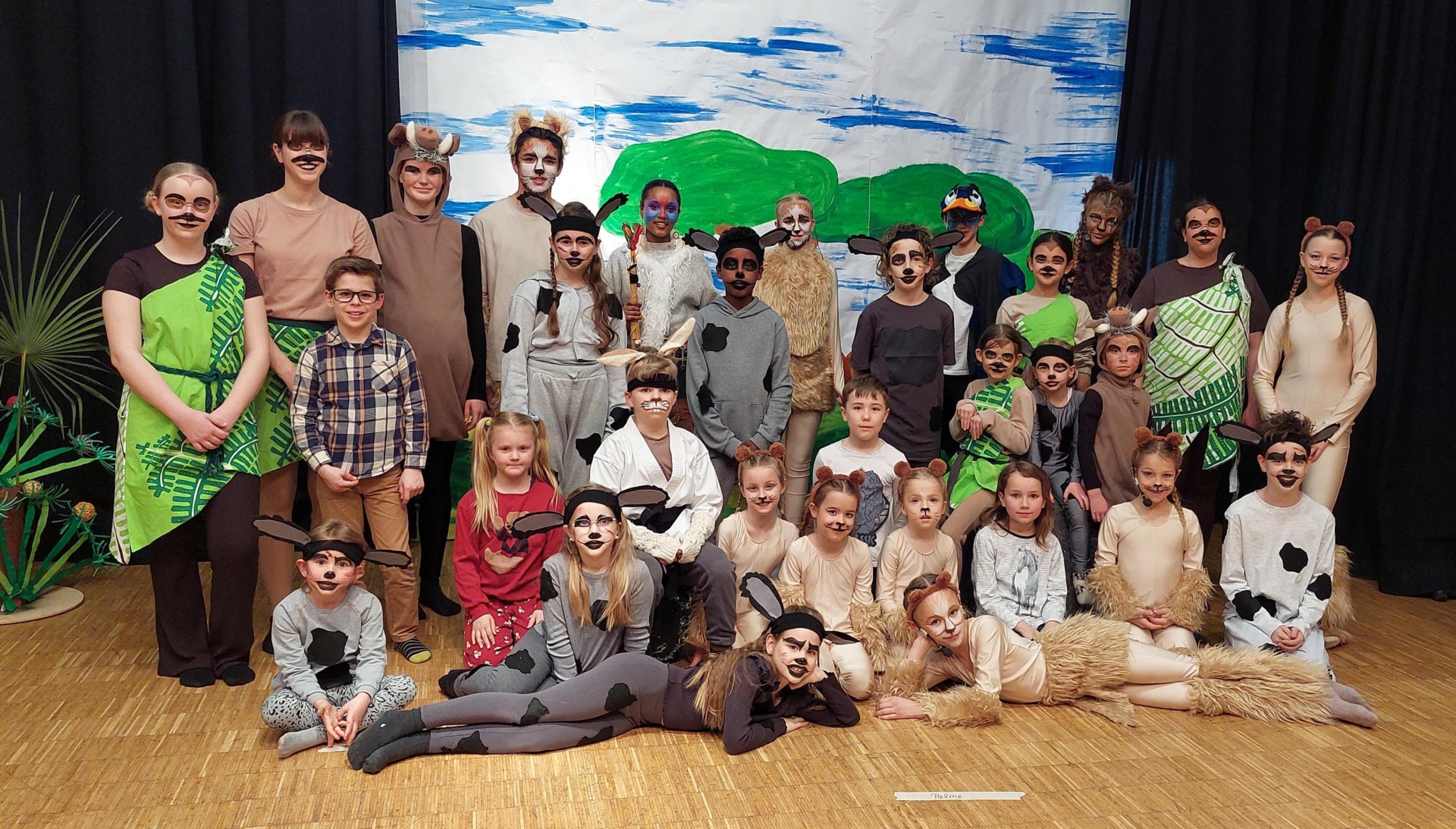 Gruppenfoto der Schauspieler des Kindertheaterstücks "Der Löwenprinz" des AHC Sielmingen: Schauspieler stehen, knien und liegen auf einem Teil der Bühne vor einer gemalten Hintergrundkulisse.