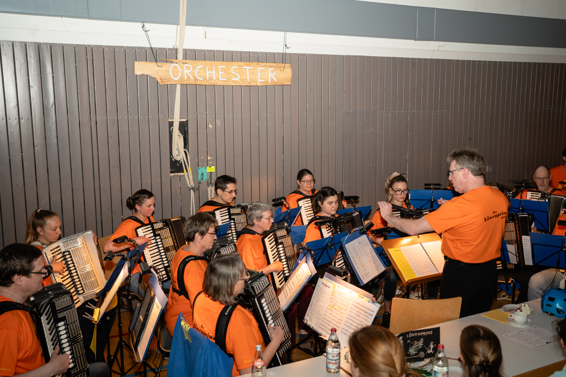 Das Orchester während der Aufführung: Zu sehen sind einige Spieler und der Dirigent Rolf Weinmann in Aktion. Sie sitzen vor einer Holzwand der Gemeindehalle Sielmingen. An der Wand hängt ein Schild mit der Aufschrift "Orchester".