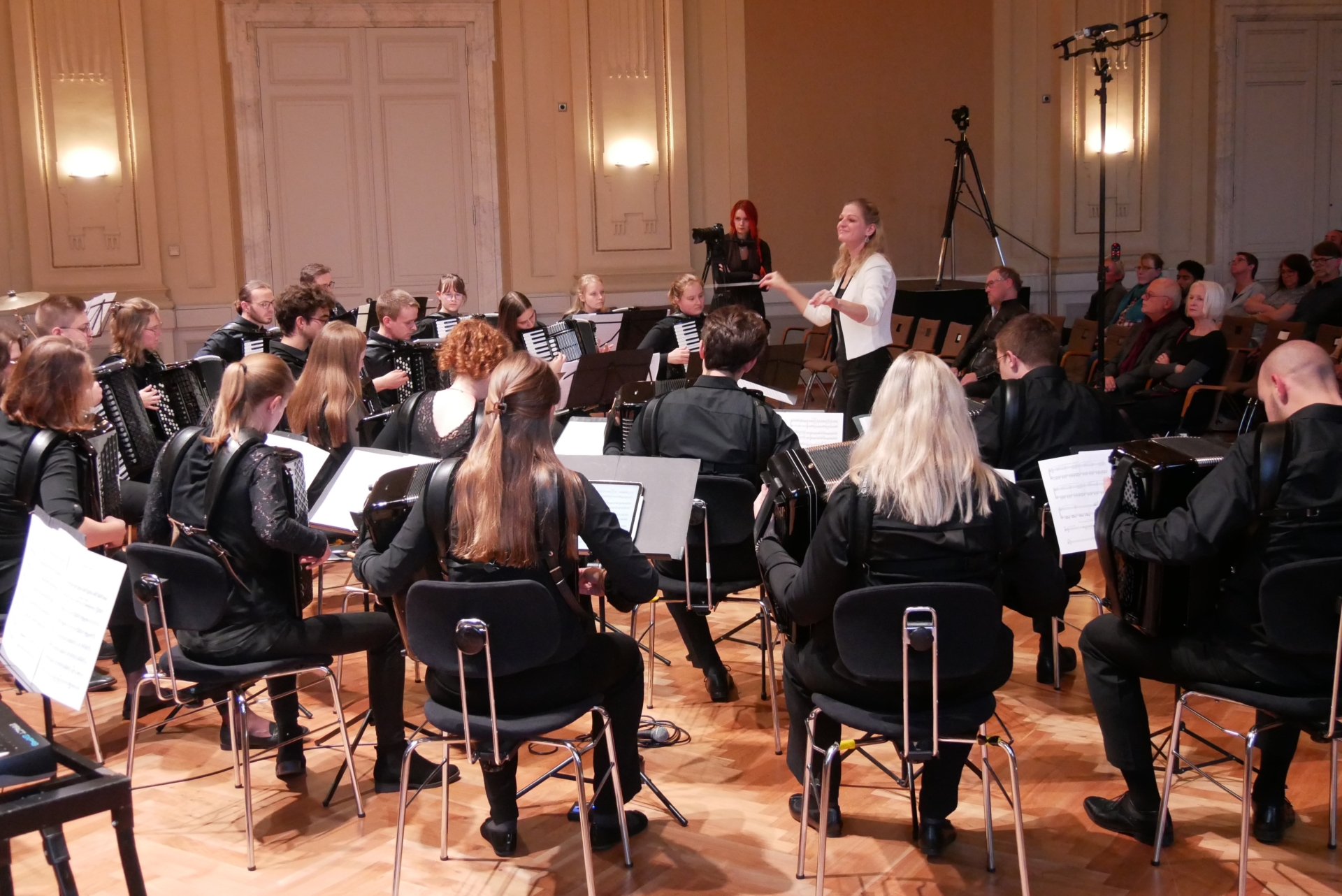 Von der linken Seite aus aufgenommenes Orchesterfoto mit Blick zur Dirigentin Silke D'Inka und dahinter dem Publikum