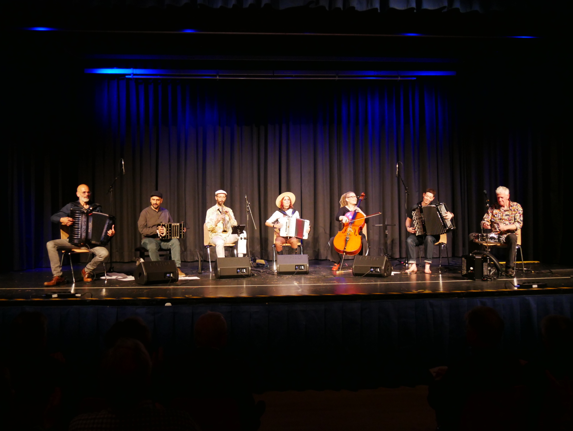 Fünf Akkordeonisten und zwei Begleitmusiker - mit Sopran-Saxophon und Cello - sitzen in einer Reihe nebeneinander auf der Bühne.